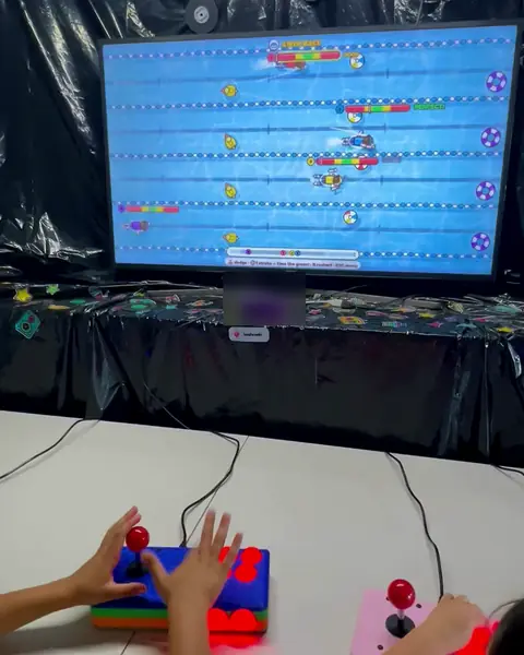 Children’s hands slapping the buttons of two arcade stations while a swimming race plays on the TV.