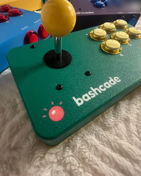 A green Bashcade arcade station with a yellow ball-top joystick and six gold buttons, with other stations behind it.