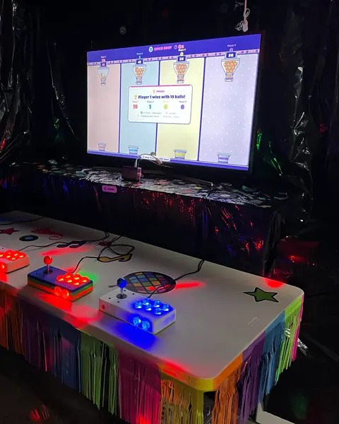 A dark garage lit by disco lights: a long table with four glowing arcade stations faces a TV showing a game’s results.
